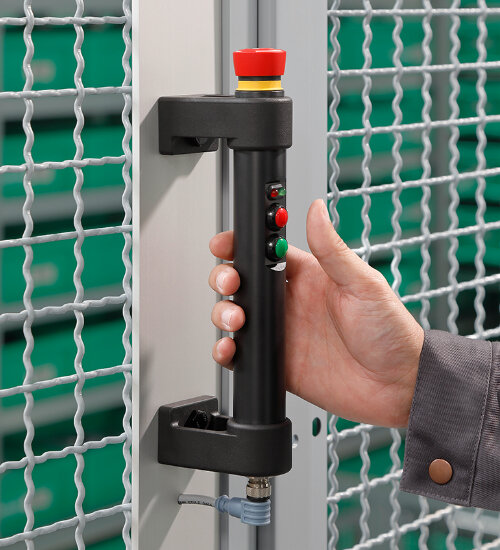 Hand gripping black industrial handle with red emergency stop button and two smaller control buttons on metal frame with wire mesh background